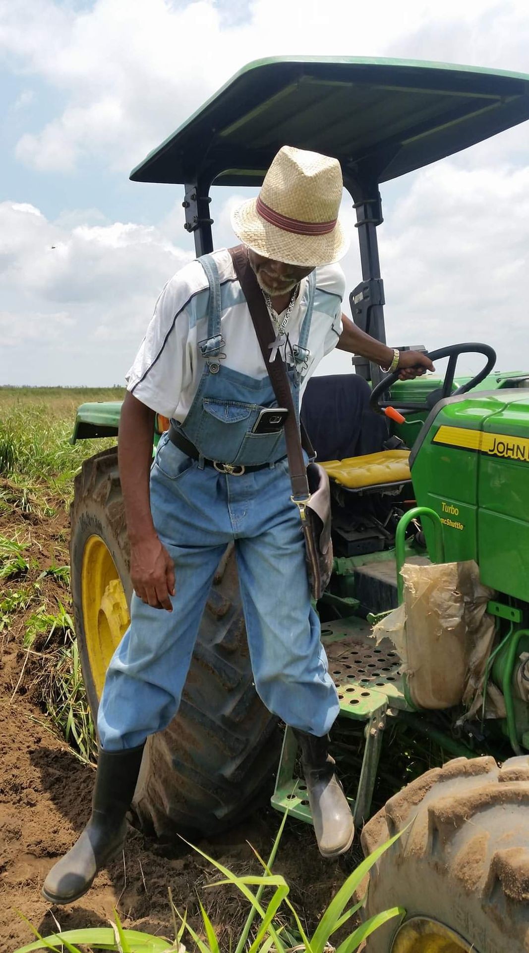 Person wearing overalls and a straw hat stepping off a green tractor in a field.