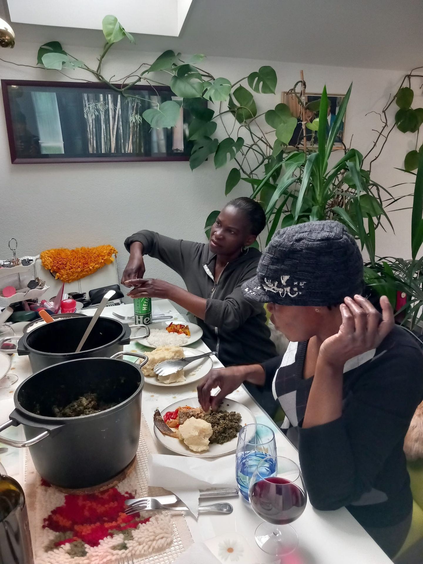 Two people sitting at a table with plates of food, surrounded by plants and kitchen items.